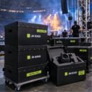 Black JM Boxes cases stacked on a dolly at an arena load-in, with a moving head light in custom foam